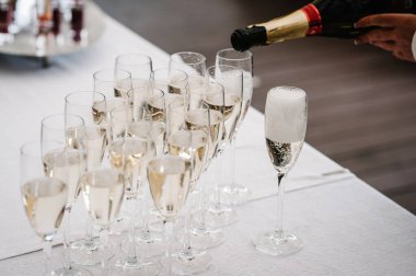 Beyaz masanın üzerinde duran cam şişeden şampanya döken erkek elleri. Kapatın. Adam kadehleri dolduruyor. Seçkin içkiler üç sıra halinde duruyor..