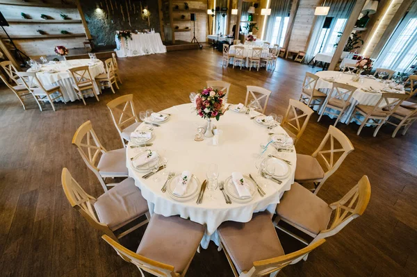 In the wedding banquet hall stand festive chairs and tables for guests covered with a tablecloth and decorated with composition of flowers and greenery. Wedding party in tent with style wooden wall.