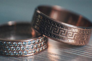 Hafif ahşap arka planda iki altın alyans. Yunan aksesuarlı erkek alyansı. Taşlı kadın yüzüğü.