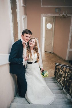 Zarif, zarif bir gelin, elini damada koydu, merdivenlerde duruyor, kapıların yanında desenler var. Kapatın. Portre. Geçmişe dönüş. Kapalı alanda klasik mimari. Sahte metal merdiven.