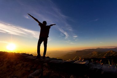 Silhouette of traveler stand exten and raise arms at top of mountain.