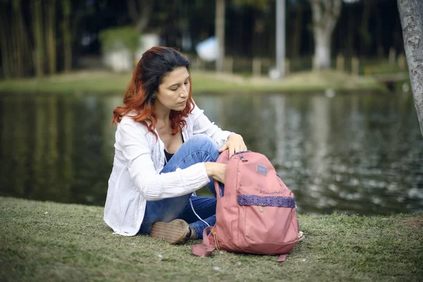 Woman sitting in the meadow looks at her pink bag. Bottom of a lake.