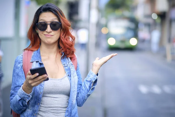 Woman makes gesture while looking at her red hair cell phone with black glasses uses her cell phone in the street.