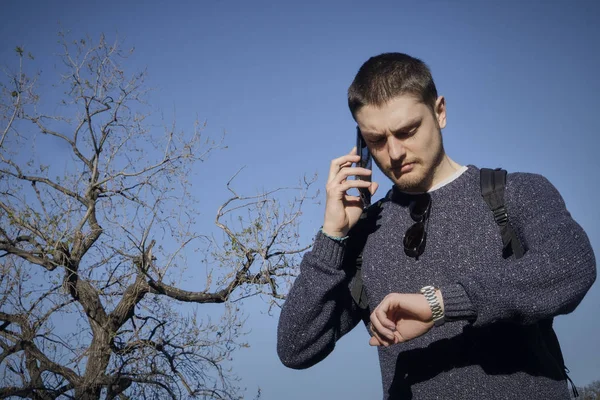 Casual young man speaks on the phone and looks at his watch. Looks very serious