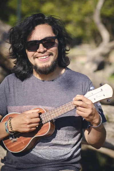 Young man sitting in a park chair playing the ukulele and looking at the camera smile