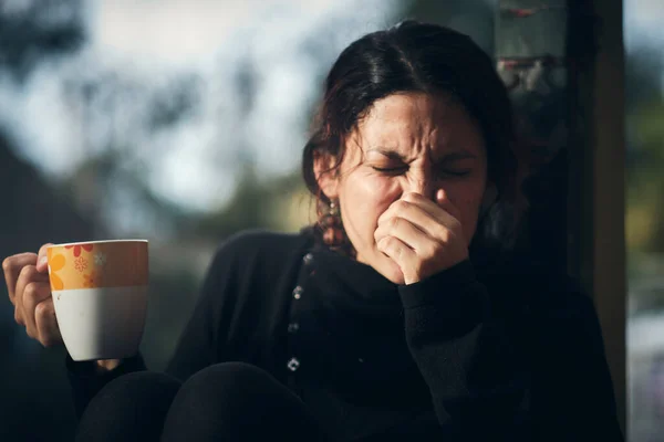 Woman sneezes hard and carries her hand to the nose, in her other hand holds a cup of tea