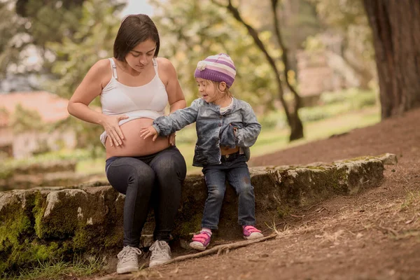 Mother and daughter sitting in a park touching the mothers pregnant belly while smiling and looking at each other