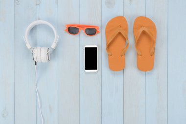 Mavi ahşap arka planda boş eşyalar: parmak arası terlikler, kulaklıklar, akıllı telefon ve güneş gözlüğü