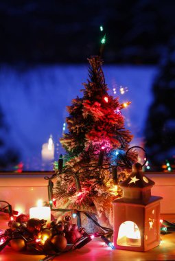 Penceresinin yanında renkli ışıklar olan Noel ağacı. Kış manzarası uzaktan görülebilir..