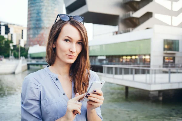 Joven sonriente mujer de negocios corredor caucásico utiliza el teléfono inteligente, mientras que de pie en la calle cerca de rascacielos . — Foto de Stock