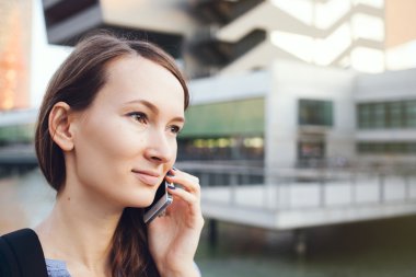 Genç gülen iyi giyimli beyaz bir iş kadın akıllı telefon gökdelenler kullanır..