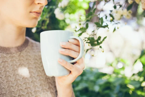 Joven mujer caucásica bebe café de la mañana de la taza azul grande siendo al aire libre en el jardín de floración de primavera . — Foto de Stock