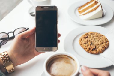 Kadının Close-Up eller holding cep telefonu ise içme kahve ve yemek tatlı - yulaf çerez Cafe.