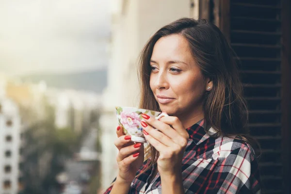 La mujer sonríe y bebe café en un balcón . — Foto de Stock