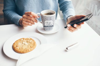 Modern kafede çay içerken ve yulaf kurabiyesi yerken cep telefonu tutan kadınların yakın çekimi. Haberler, sosyal ağlar, akıllı telefondan internette sörf yapmak. Çay molası işi
