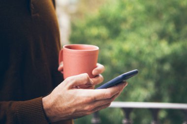 Seramik bardaktan sıcak içecek içerken cep telefonu tutan adamın ellerini yakın çekim. Haberler, e-postalar, makaleler, sosyal ağlar, akıllı telefondaki flört uygulamaları. Bacony, yeşil ağaçların manzarası