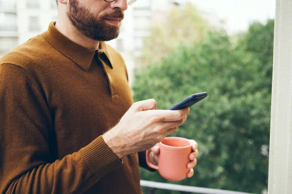 Handsom beard man in sweater is using mobile phone and drinking coffee while standing on the balcony with green trees view. Technology and applications concept, online food ordering.