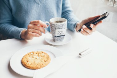 Kadın çay içiyor ve modern bir kafede kahvaltı yapıyor. Kız cep telefonu tutuyor, sosyal ağlarda güzel sohbetlerden hoşlanıyor, internette komik makaleler okuyor, mavi kazak giyiyor..