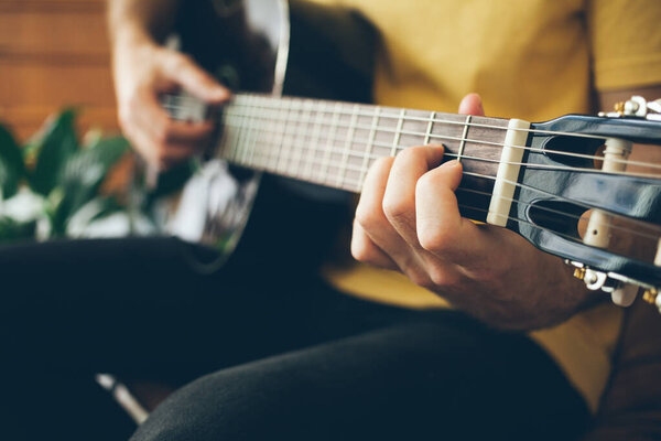 Close-up of a man hands playing black color guitar at home in beautiful modern interior. Natural light and selective focus. Guitarist in yellow t-shirt.