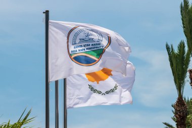 Agia Napa belediyesi bayrağı ve Kıbrıs bayrağı. Bayrak direğinde ve sokak lambasında bayraklar var. Rüzgar bayrağı şişiriyor.
