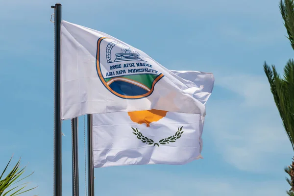 Agia Napa belediyesi bayrağı ve Kıbrıs bayrağı. Bayrak direğinde ve sokak lambasında bayraklar var. Rüzgar bayrağı şişiriyor.