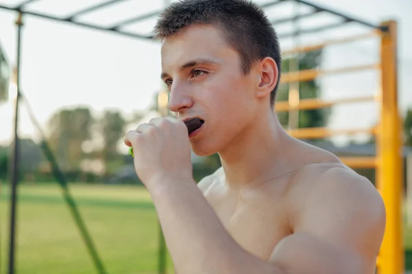 Man is eating carbohydrate bar during workout. Outdoor gym backg — Stock Photo, Image