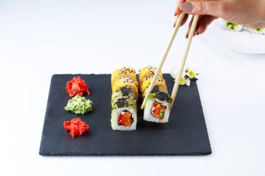 Suşi seti zencefil ve wassabi ile siyah kare bir tabakta hafif bir zemin üzerinde. Kadın eli yemek çubuklarıyla suşi tutuyor. Yakın çekimler