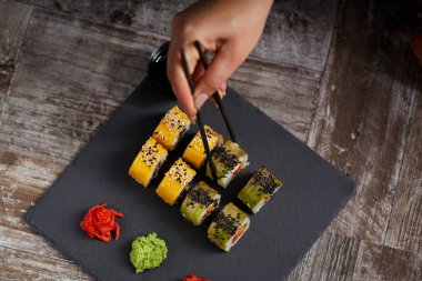 Suşi kare siyah bir taş tabakta. Dişi bir el bir rulo çubuk tutuyor. Yukarıdan bak. Eski Orman Arkaplanı