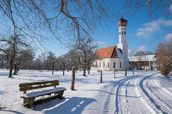 Idyllic village church, wintry road and wooden bench — Stock Photo, Image