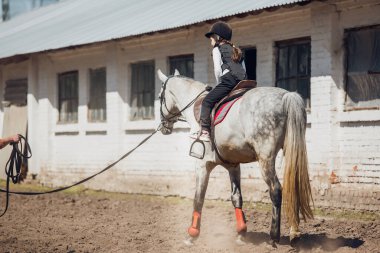 Kasklı küçük kız ata binmeyi öğreniyor. Eğitmen çocuğa binicilik öğretiyor..