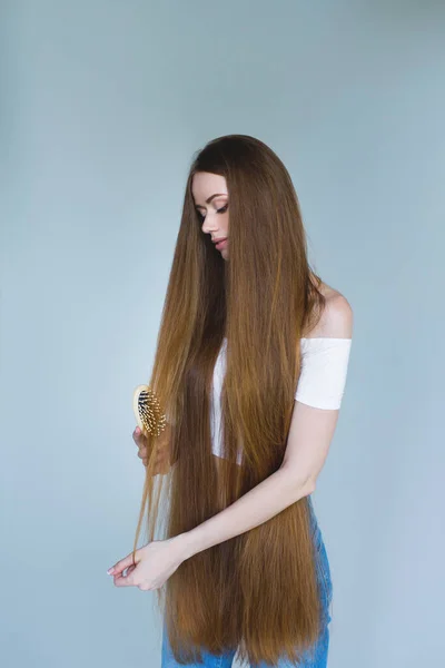 Concept of hair loss. Close up portrait of unhappy sad stressed young woman with long dry brown hair, she is looking at the brush in hand, isolated on grey background. — Stock Photo, Image