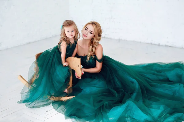 Porträt einer schönen jungen Mutter mit einer niedlichen blonden Tochter im weißen Interieur in eleganten grünen Kleidern. — Stockfoto