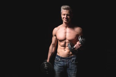 Dambılı olan yakışıklı atletik adam kendinden emin bir şekilde ileriye bakıyor. Güçlü vücut geliştirici, karın kasları, omuzları, pazıları, trisepsleri ve göğsü var. Kuvvet ve motivasyon