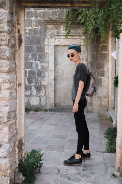 Sırt çantalı siyah güneş gözlüklü genç bir kız taş duvarlarla çevrili bir kaldırım taşı sokakta duruyor..