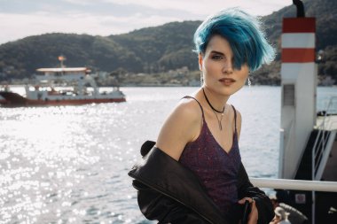 Mor elbiseli, mavi saçlı ve siyah ceketli bir kızın portresi dağlardaki bir körfezin arka planında