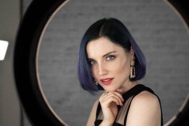 Profesyonel makyajlı, mavi saçlı ve mükemmel ciltli genç bir kız stüdyoda sosyal medya için poz veren bir lambanın önünde oturuyor. Güzellik blogu. Boşluğu kopyala.