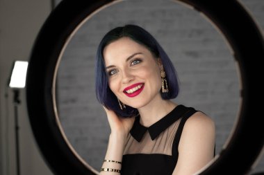 Profesyonel makyajlı, mavi saçlı ve mükemmel ciltli genç bir kız stüdyoda sosyal medya için poz veren bir lambanın önünde oturuyor. Güzellik blogu. Boşluğu kopyala.