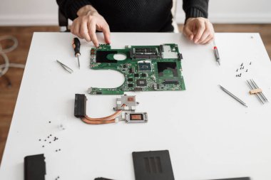 Beyaz bir masada oturan bilgisayar tamircisi. Masanın üzerinde parçalanmış bir bilgisayar veya dizüstü bilgisayarın farklı parçaları var. Ana kart, soğutma sistemi, vida ve tornavida.