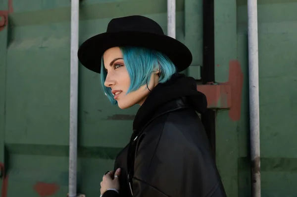 一个头戴黑色帽子 身穿皮夹克 头戴蓝色头发的女孩站在一个绿色的容器旁边 免版税图库照片