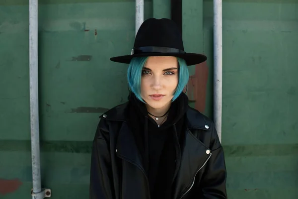 一个头戴黑色帽子 身穿皮夹克 头戴蓝色头发的女孩站在一个绿色的容器旁边 图库图片