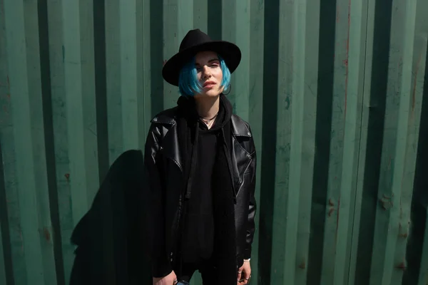 一个头戴黑色帽子 身穿皮夹克 头戴蓝色头发的女孩站在一个绿色的容器旁边 复制空间 免版税图库图片