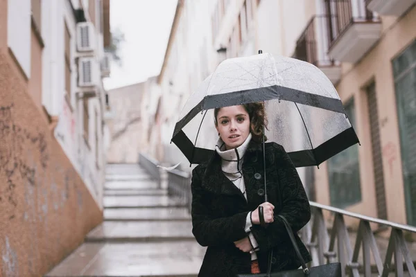 Mujer joven con rostro frío camina por las calles de Cáceres con un paraguas transparente en un día lluvioso — Foto de Stock