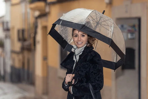 Sötét hajú fiatal nő sétál Caceres utcáin egy átlátszó esernyővel egy esős napon. — Stock Fotó