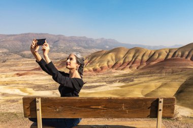 Gri iplikli genç bir esmer boyalı tepelerdeki bankta selfie çekiyor.