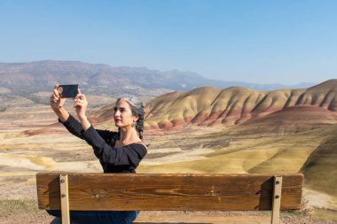 Gri iplikli genç bir esmer boyalı tepelerdeki bankta selfie çekiyor.