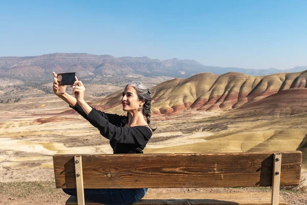 Egy fiatal barna hajú, szürke szálakkal, aki szelfiket készít egy padon ülve a Painted Hills-i kilátásban. — Stock Fotó