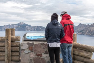 Bir çift, arka planda Krater Gölü manzaralı bir bilgi paneline bakıyor.