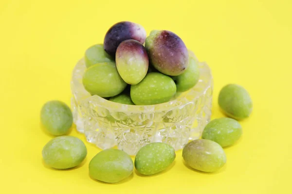 Přírodní čerstvé kulaté zelené a hnědé olivy v dekorativním skleněném talíři na žlutém pozadí — Stock fotografie