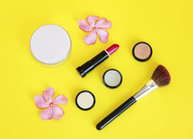 Dekoratif kozmetik ürünleri, pudra, göz farı, ruj, dudak parlatıcısı, makyaj fırçası ve pembe çiçekler.