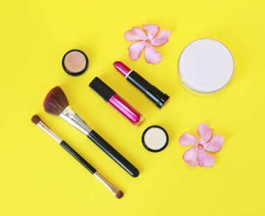 Dekoratif kozmetik ürünleri, pudra, göz farı, ruj, dudak parlatıcısı, makyaj fırçası ve pembe çiçekler.